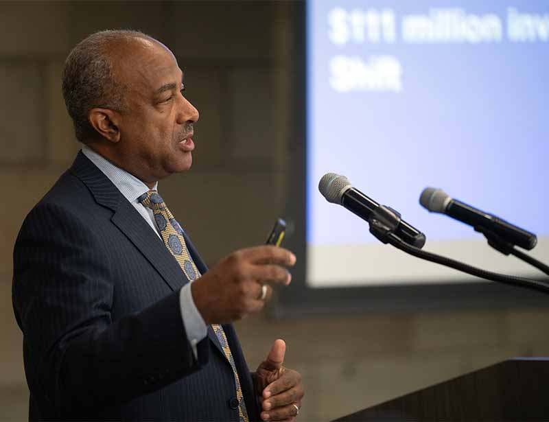 Chancellor Gary S. May speaks, with PowerPoint slides visible in background