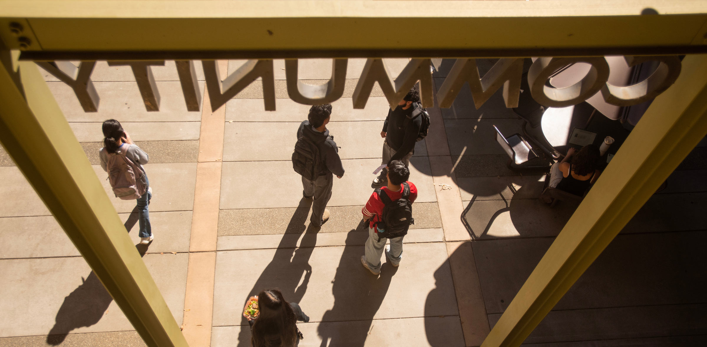 Overhead photo of pedestrians and long shadows on a sunlit sidewalk framed by yellow window