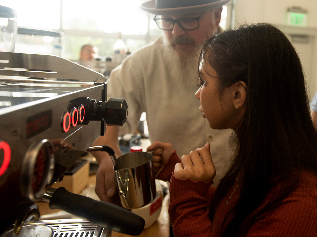 A student in a burgundy long-sleeve shirt carefully uses the milk frother on an espresso machine while being supervised by a man with a white beard, wearing square-rimmed glasses and a fedora.