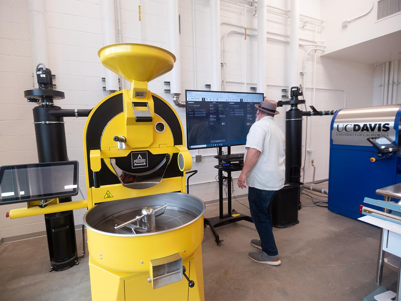 A man in a white, short-sleeved, button-up shirt, dark blue jeans, gray sneakers and a feathered gray fedora uses a computer with a large monitor between two large coffee roasters branded in UC Davis' Aggie Blue and Aggie Gold colors.