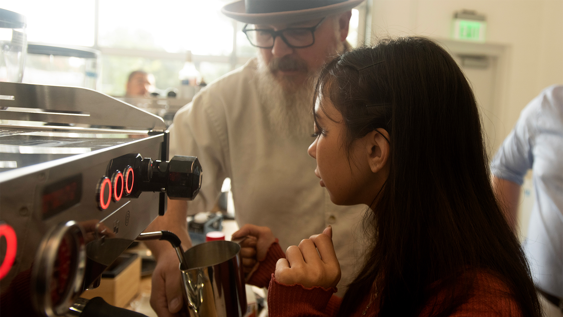 A student in a burgundy long-sleeve shirt carefully uses the milk frother on an espresso machine while being supervised by a man with a white beard, wearing square-rimmed glasses and a fedora.