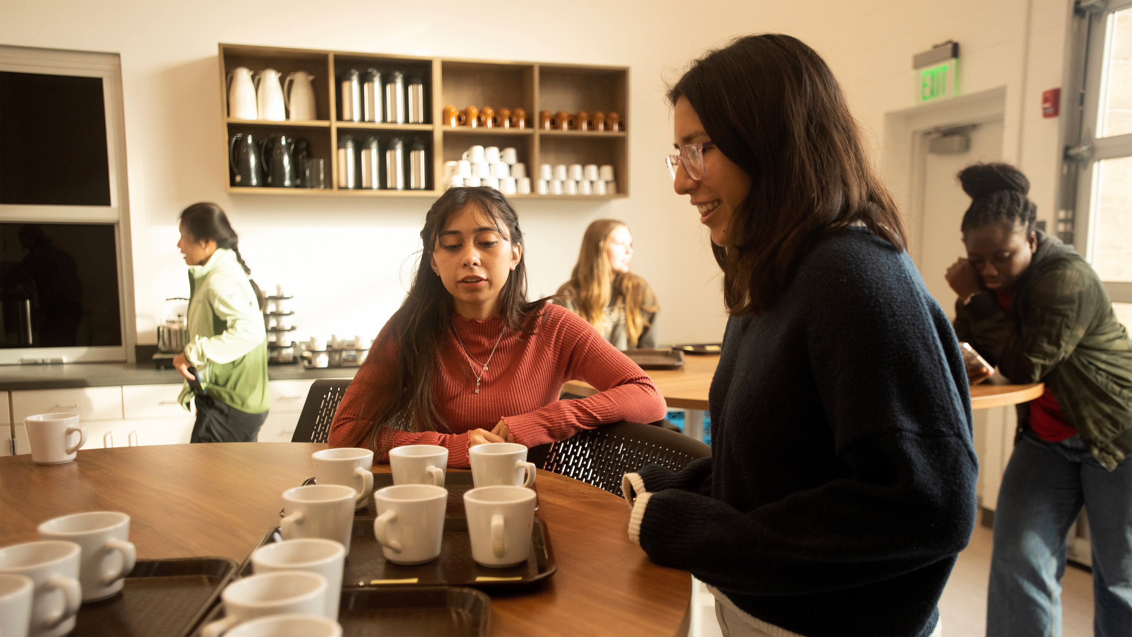 Two students talk at a wooden table with trays of white coffee mugs while other students mingle and check their phones behind them.