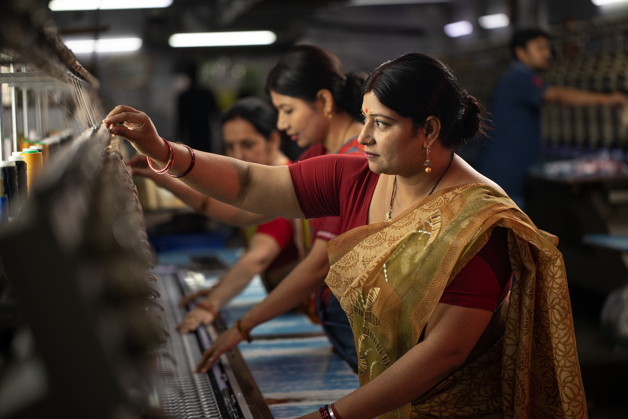 Three women operating textile machines in a dim factory, one wearing a gold sari