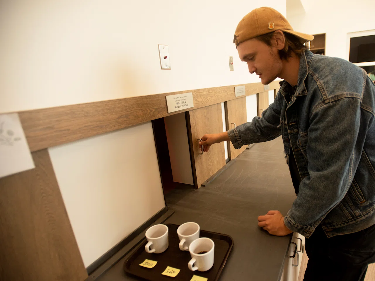 Keegan Thompson, in a backward mustard baseball cap and blue denim jacket, slides open a wooden service door in a white wall, about to deliver the tray with three filled coffee mugs on the dark countertop beside him.