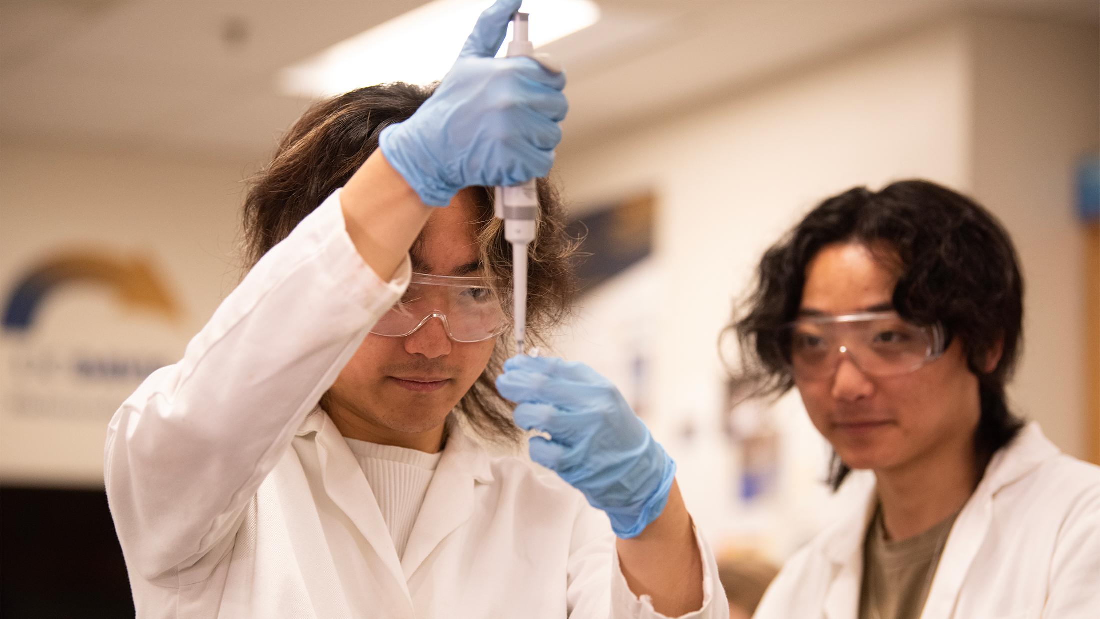 Two students wearing white lab coats, blue gloves and safety goggles work together in a laboratory. One student is using a pipette to transfer liquid, while the other observes attentively.