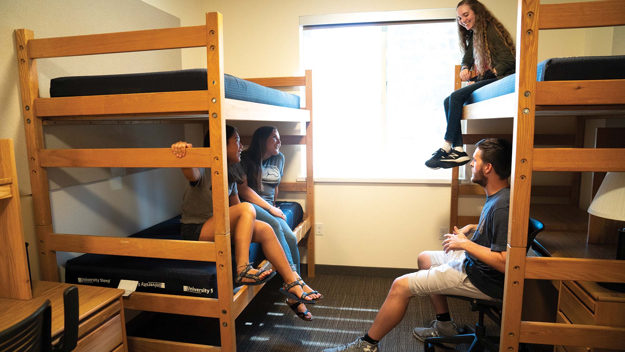 Four students sit talking on bunk beds in a furnished, but unoccupied, dorm room. Desks, lamps and chairs are also seen.