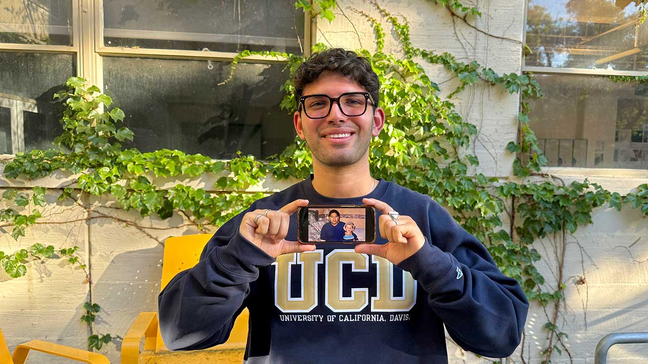 Standing outside, a smiling man holds up to his chest an iPhone to show a photo of himself as a child with his fatherh
