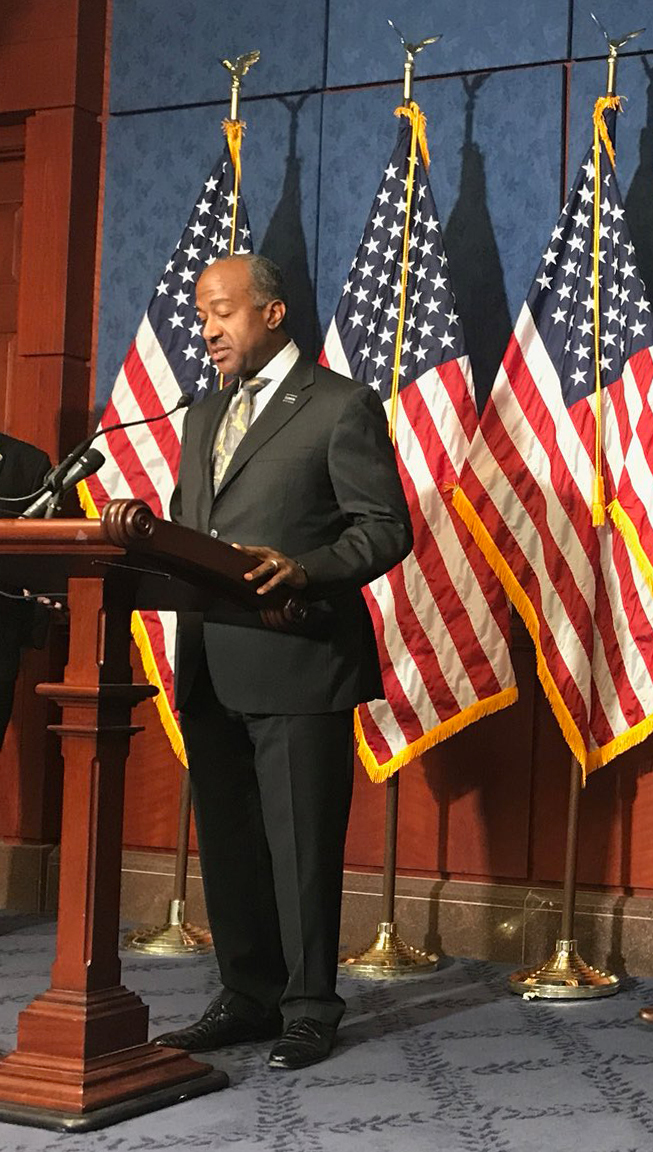 Chancellor May at podium, with backdrop of U.S. flags