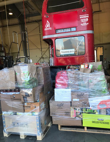 Food on pallets, behind vintage double-decker bus from London.