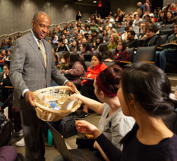 Chancellor May passes out cookies, from large basket, in lecture hall.