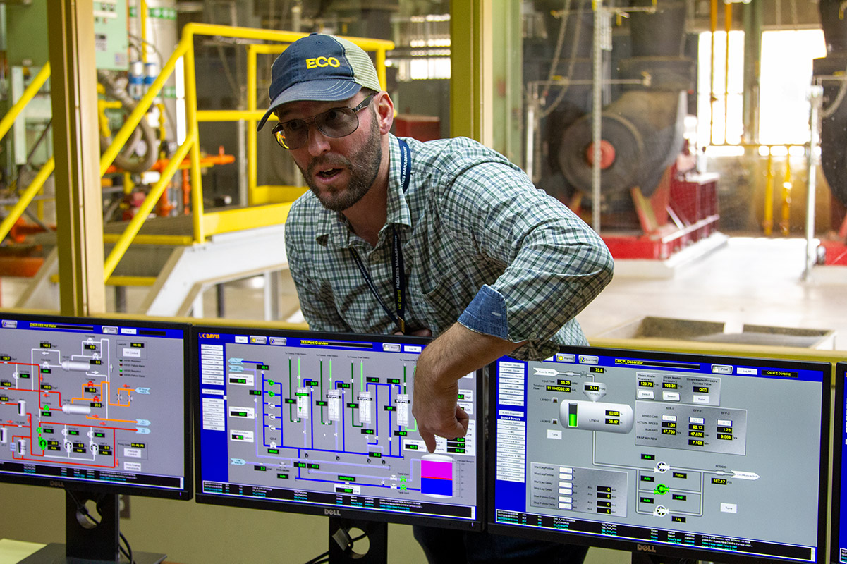 A man wearing glasses and a hat points to computer screens.