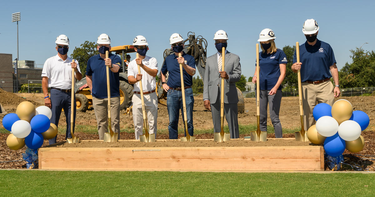 Seven people dig into a trough of dirt, with golden shovels.