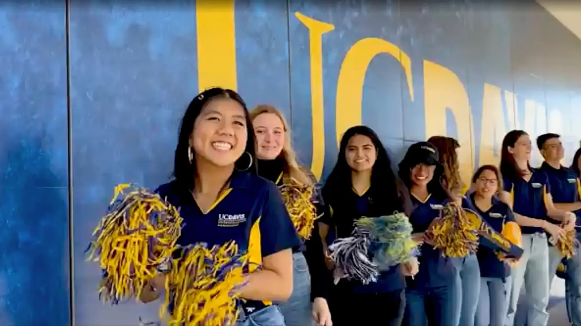 Students with pom poms gathered in front of a wall that reads, UC Davis, to cheer on new Aggies. 