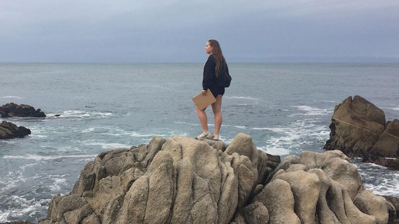 A woman stands on giant shoreline rocks looking out at the ocean and holding a clipboard