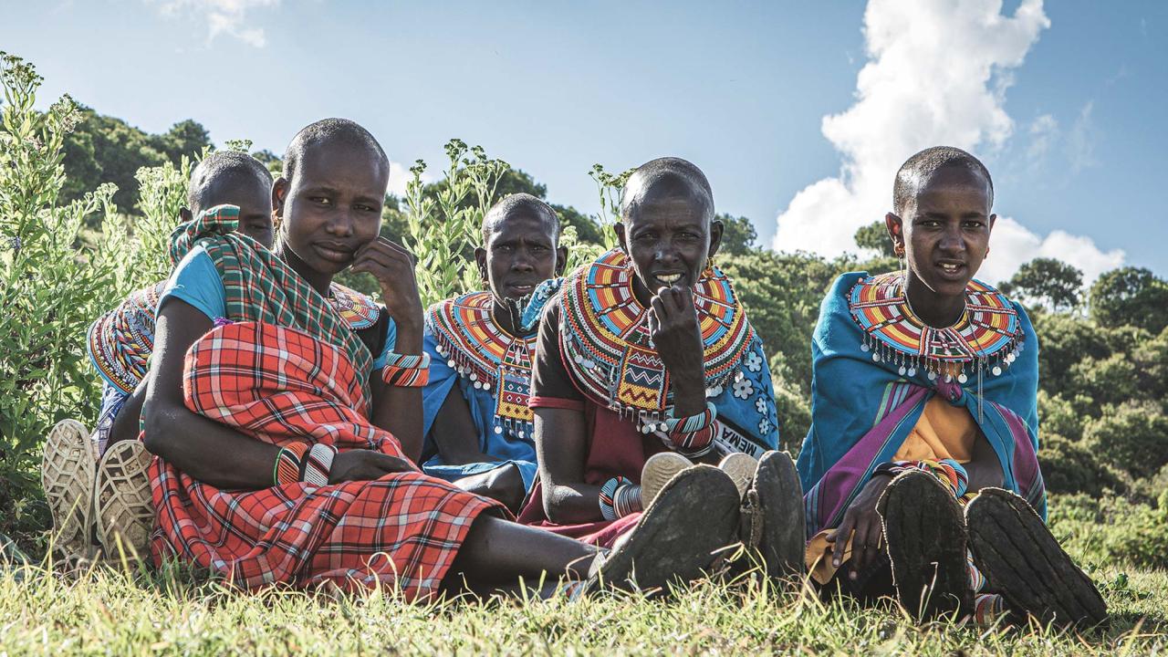 Low-angle photo of five Kenyan women sitting on the ground. They are dressed in colorful traditional clothes and looking at the camera.