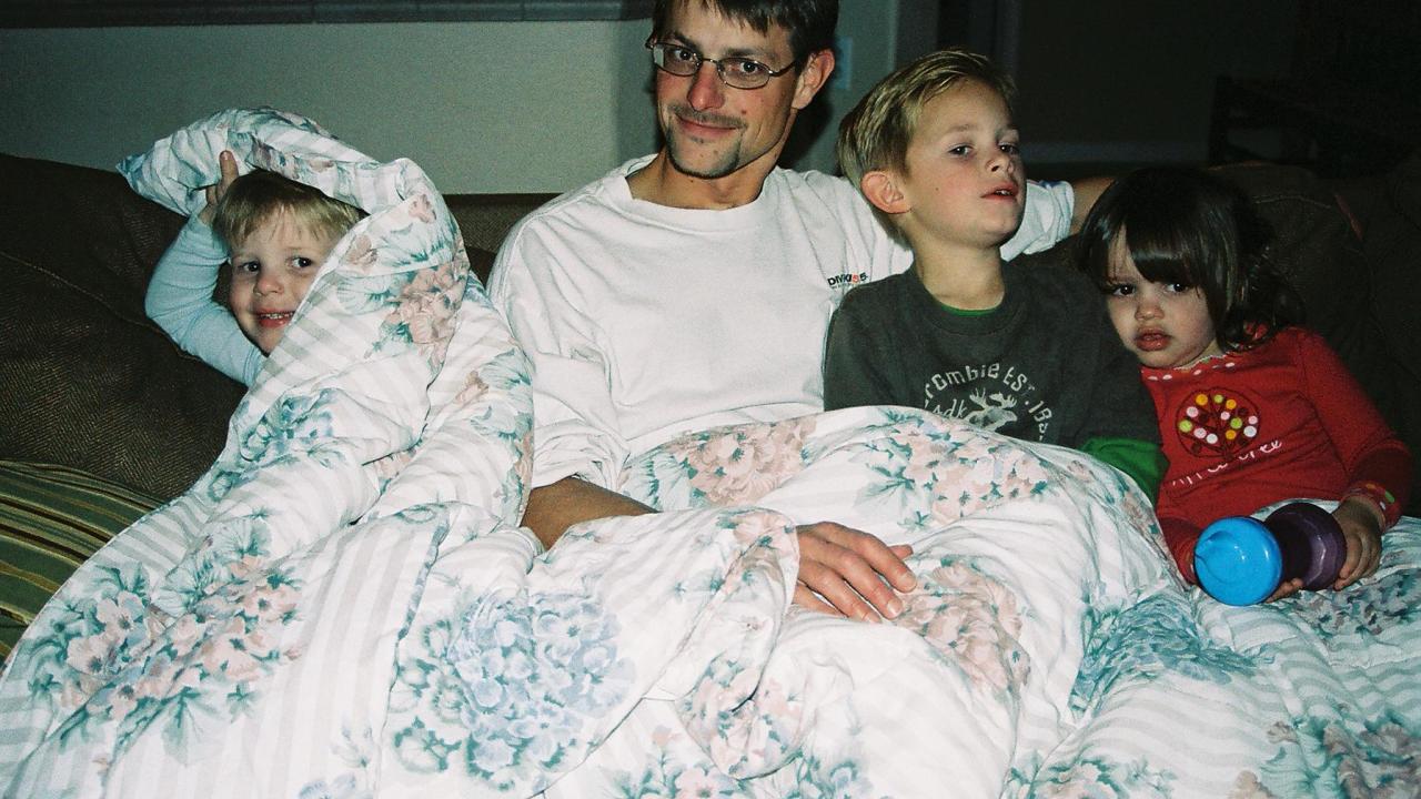 One adult in a white shirt and three kids, one in a blue shirt, one in a gray shirt and another in a red shirt, sit together on a brown couch under a white flower-patterned blanket.