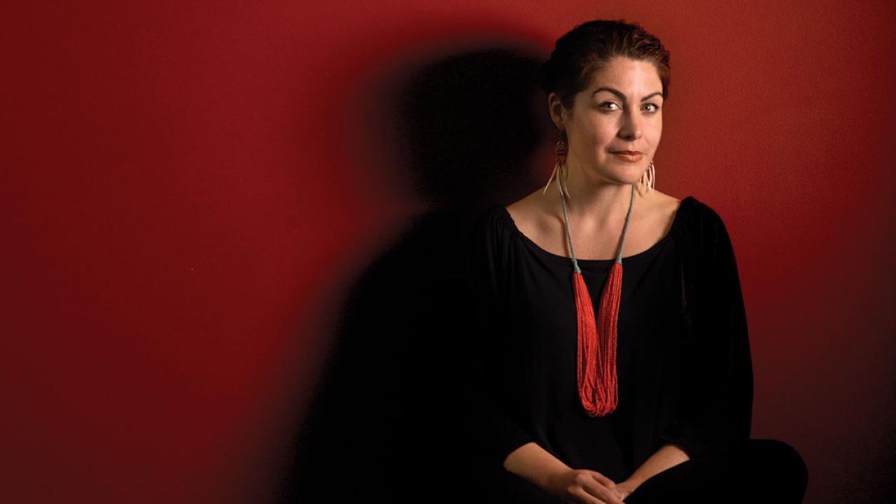 Portrait of a woman looking at the camera. She is wearing a black shirt and red necklace, standing against a red wall. She is seen from the waist up.