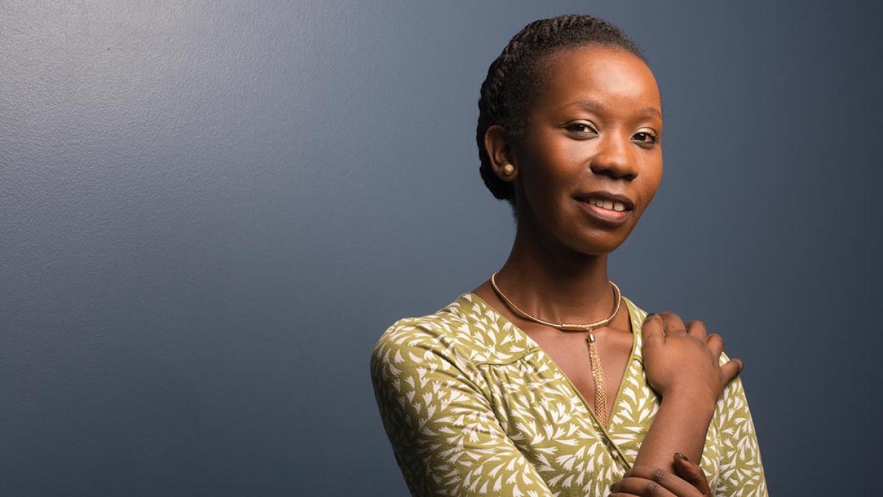 Portrait of a woman against a blue wall. She looks at the camera and has her right hand resting on her left shoulder.