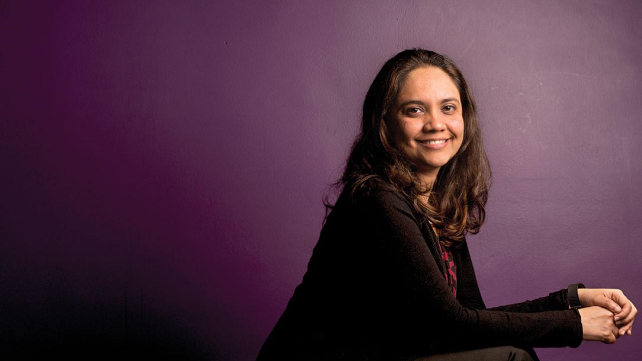 Portrait of a woman, seated and looking at the camera. She is turned to her left, and seen from the elbows up. She wears a black shirt and is in front of a purple wall.
