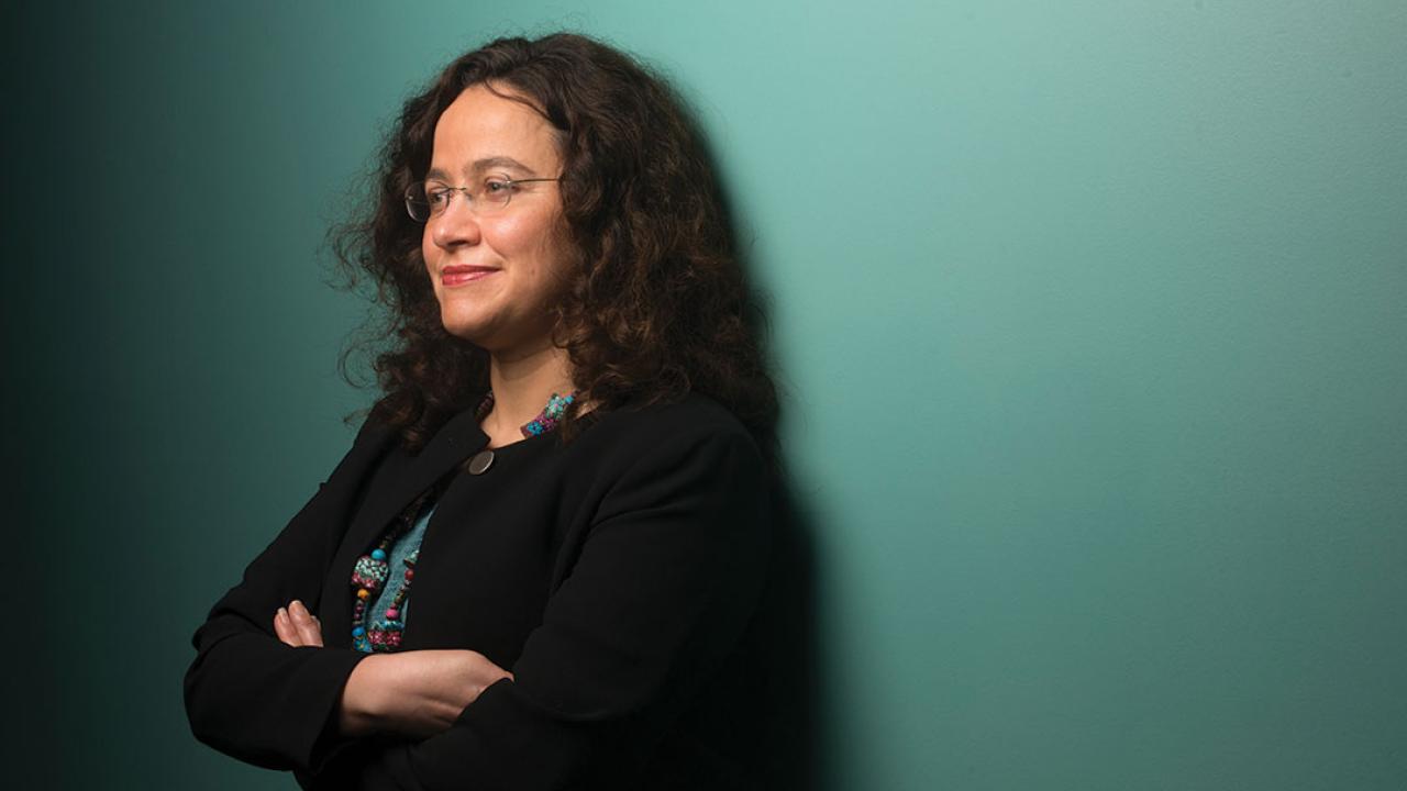 Photo of a woman leaning against a green wall and looking off camera to the left. She standard with her arms crossed, wears a black short with colorful necklace and wears glasses.