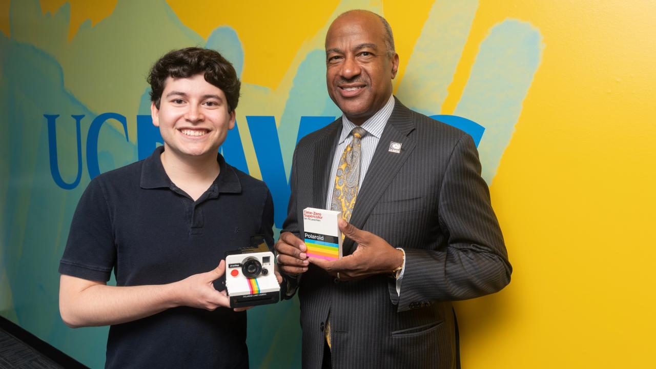 A young man in a black polo shirt holds a Polaroid camera made out of Legos against his chest. He is standing to the left next to Chancellor Gary S. May who is wearing a dark suit and holding a small box of Polaroid film. Both are smiling at the camera in a studio setting with a bright background that has blue UC Davis logo along a yellow wall.