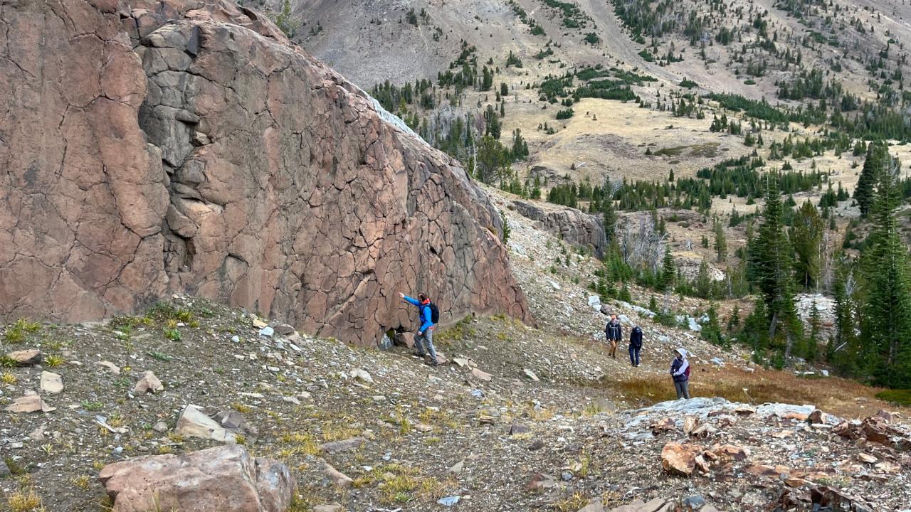 Outdoor scene of a cliff of rock with mountains in the background. Small human figures can be seen at the base of the cliff. 