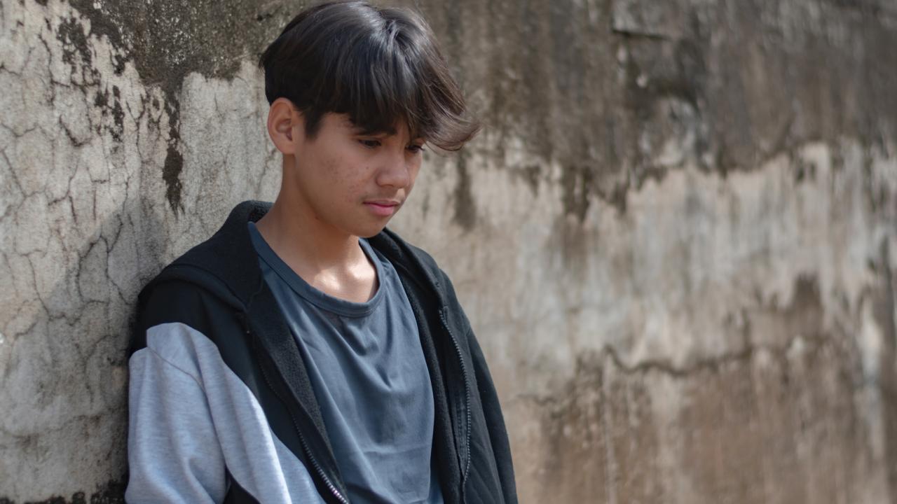 A boy with dark hair leans against a textured wall, looking down thoughtfully.