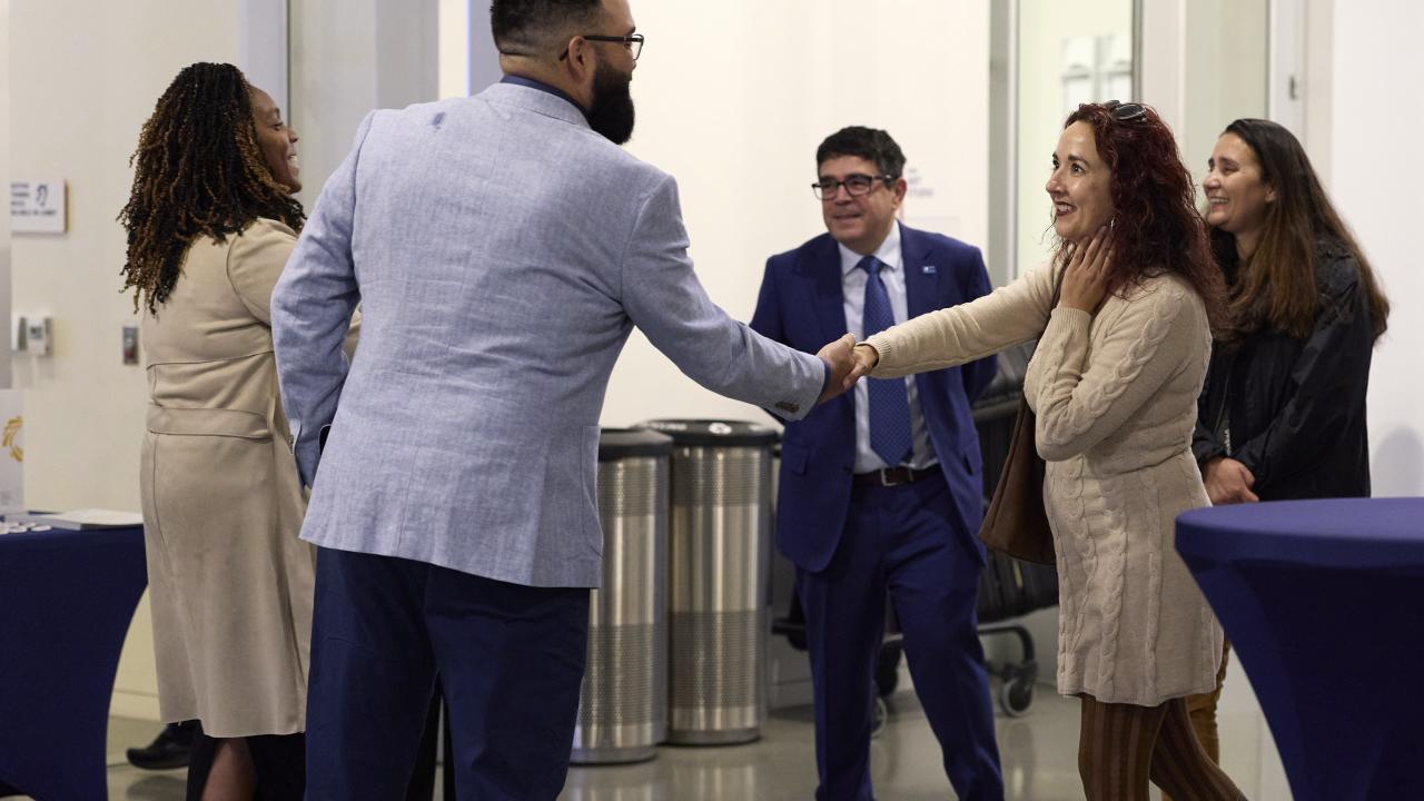 in a mixer setting, academics in professional dress introduce each other by shaking hands. 