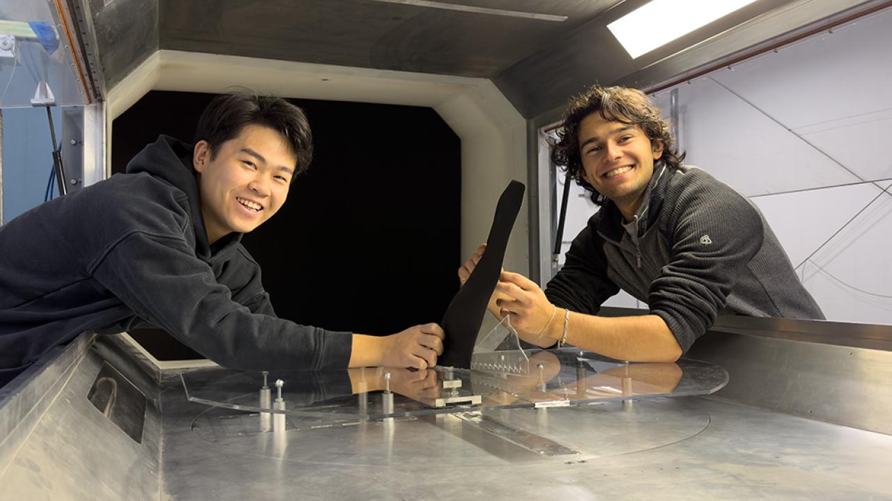 Two young men in dark clothing lean over a bench and turn to face the camera. Between them is a black fin-like object. 
