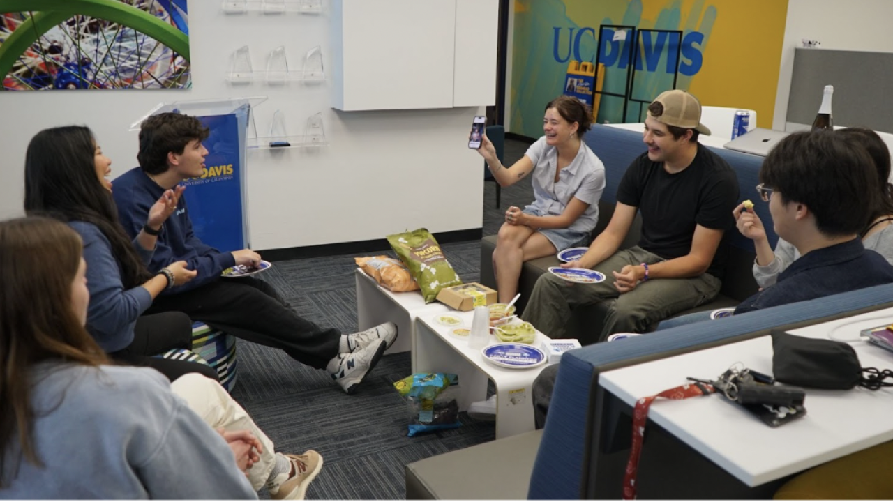 Seven students sit in a circle, laugh and eat snacks together in a UC Davis lounge. 