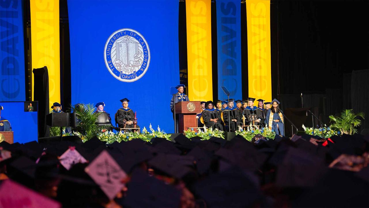Graduation stage with blue-and-gold backdrop, university seal, speakers and mortarboards