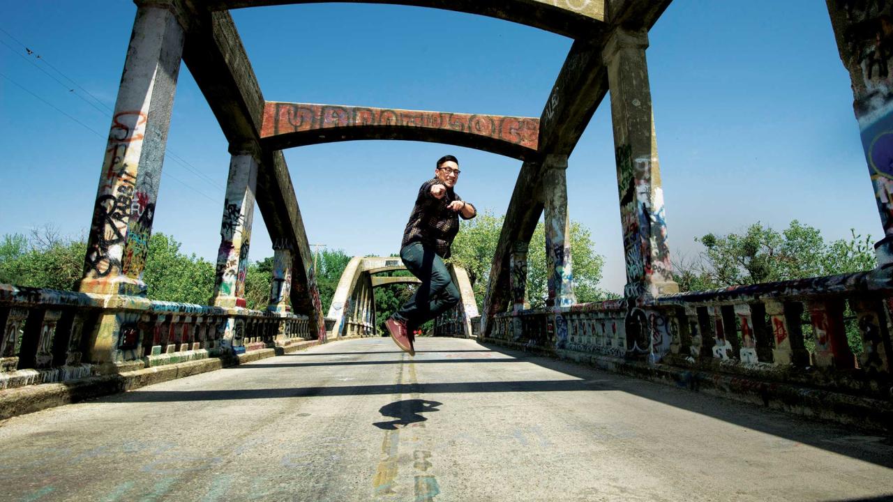 A man jumps in the air in the middle of a road that's on a bridge. The bridge is an old two-lane bridge with concrete trusses overhead. It's covered in graffiti.