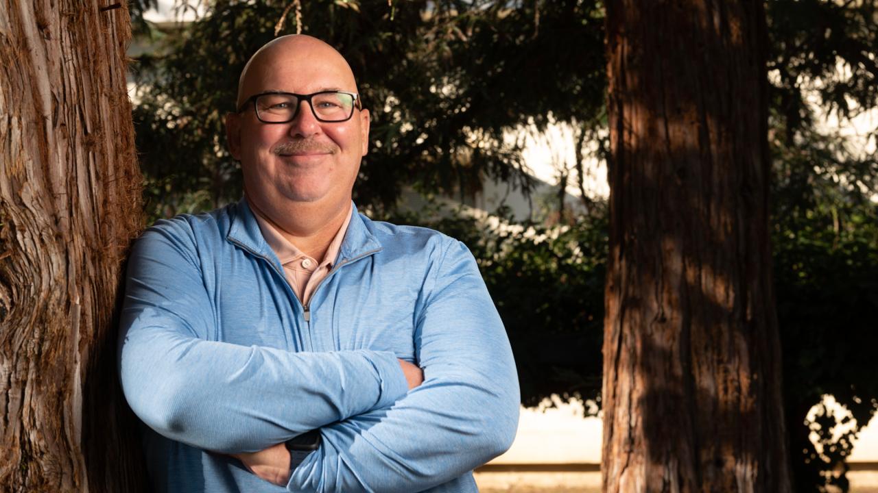 Smiling man with glasses stands confidently against a tree, arms crossed.