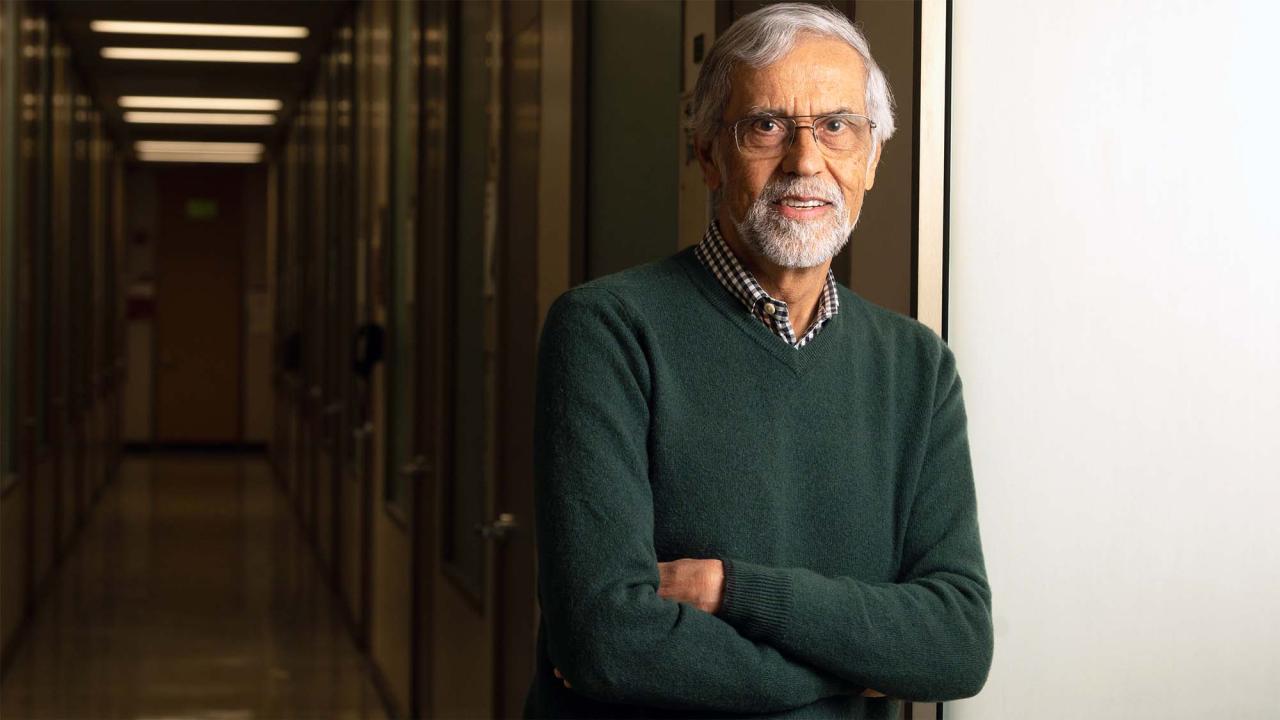 A man with glasses and a beard smiles while standing in a hallway with office doors.