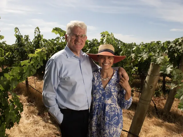 Portrait of a couple standing in a vineyard on a sunny day. The man wears a light colored shirt and glasses. The woman wears a sundress and large-brimmed straw hat.