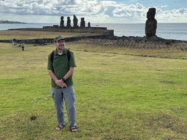 A man stands on a grassy field a row of large stone statues and the ocean behind him.