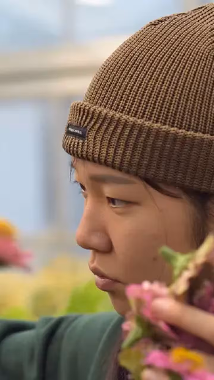 Person in brown beanie tending pink and yellow flowers by a window, photograph