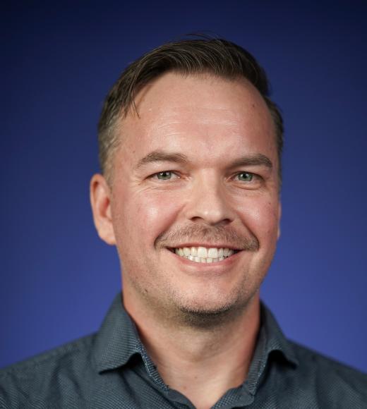 Smiling man with short hair wearing a dark blue button-up shirt, set against a purple background.