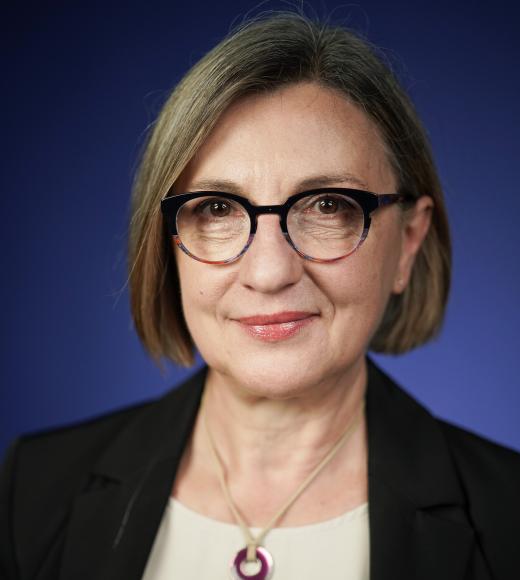 Woman with short gray hair and glasses, wearing a black blazer and colorful necklace, smiling.