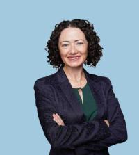 Smiling curly-haired professional woman in blazer, arms crossed, pale blue background (photo)