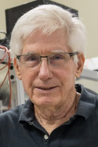 Older man with white hair and glasses, slight smile, dark polo, lab equipment in background