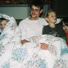 One adult in a white shirt and three kids, one in a blue shirt, one in a gray shirt and another in a red shirt, sit together on a brown couch under a white flower-patterned blanket.