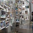 On a rainy day, a student in a black parka opens a glass door and enters a museum lobby. The glass door and its nearby wall are covered with photographs of UC Davis students standing next to the Egghead sculptures created by Robert Arneson.