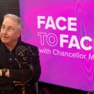 Inside a recording studio, in front of a purple backdrop with the logo for Face to Face with Chancellor Gary S. May sits a man with his arms crossed wearing glasses and short grey hair, staring to their right. 