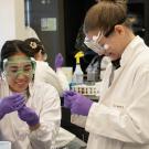 Two students in white lab coats, safety goggles and gloves work with small tubes