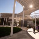 Sunlit modern open-air courtyard with curved wooden canopy, white columns, and two people walking