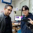 At left, a man with short curly dark hair wearing a grey jacket looks at a device held up by a younger man wearing a dark blue hoodie and glasses. 