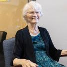Smiling woman with white hair seated in wooden chair, teal dress