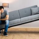 A despondent man sits at one end of a sofa wedged in a space too narrow for it. 