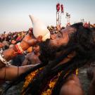 Man participates in festival in India, blowing into conch shell, while others look on 